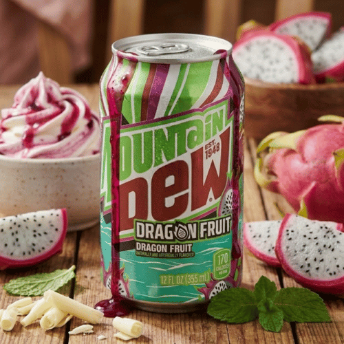 Eine grün-pinke Dose Mountain Dew Dragon Fruit steht auf einem rustikalen Holztisch. Die Szene ist dekoriert mit aufgeschnittenen Drachenfrüchten, Minzblättern und weißer Schokolade. Im Hintergrund ein Dessert mit Beeren-Topping.