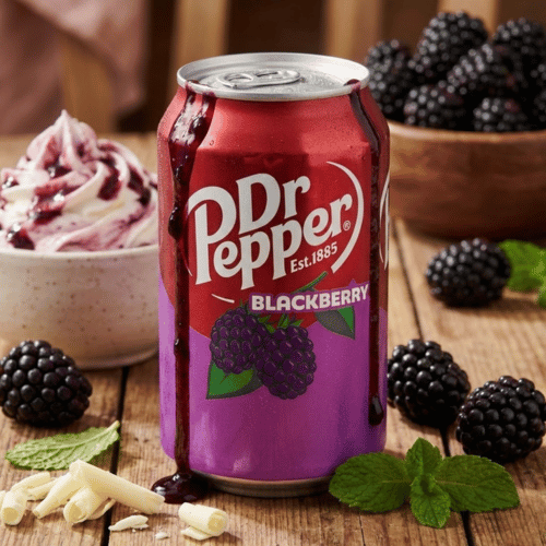 Eine rötlich-violette Dose Dr Pepper Blackberry steht zentral auf einem rustikalen Holztisch. Die Szene ist dekoriert mit frischen, dunklen Brombeeren, grünen Minzblättern, weißen Schokoladenraspeln und einem cremigen Beeren-Dessert im Hintergrund.