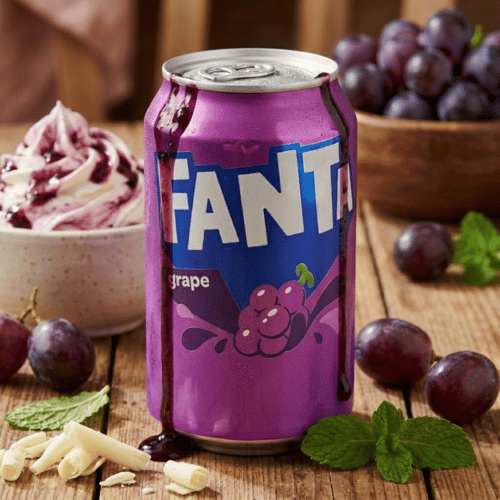 Eine violette Dose Fanta Grape steht auf einem rustikalen Holztisch. Die Szene ist umgeben von frischen, dunklen Weintrauben, Minzblättern und weißer Schokolade. Im Hintergrund ein cremiges Dessert mit lila Topping.
