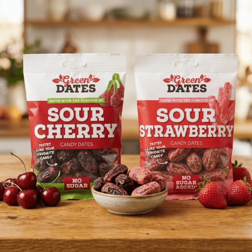 Zwei stehende Beutel Green Dates Candy Datteln in den Sorten Sour Cherry und Sour Strawberry auf einem rustikalen Holztisch, liebevoll dekoriert mit frischen Kirschen, reifen Erdbeeren und einer Schale naturbelassener Datteln.