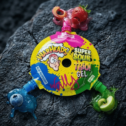 Dynamische Produktinszenierung des Warheads Super Sour Trio! Gels auf dunklem Naturstein: Aus drei farbigen Nozzles fließen leuchtend rote, blaue und grüne Gel-Texturen, die von einer frischen Erdbeere, Blaubeere und einem Apfelstück umrahmt werden – visuelle Darstellung des extrem sauren Geschmackserlebnisses.